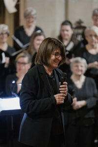 JoAnne Wasserman in front of the Santa Barbara Choral Society members holding a microphone